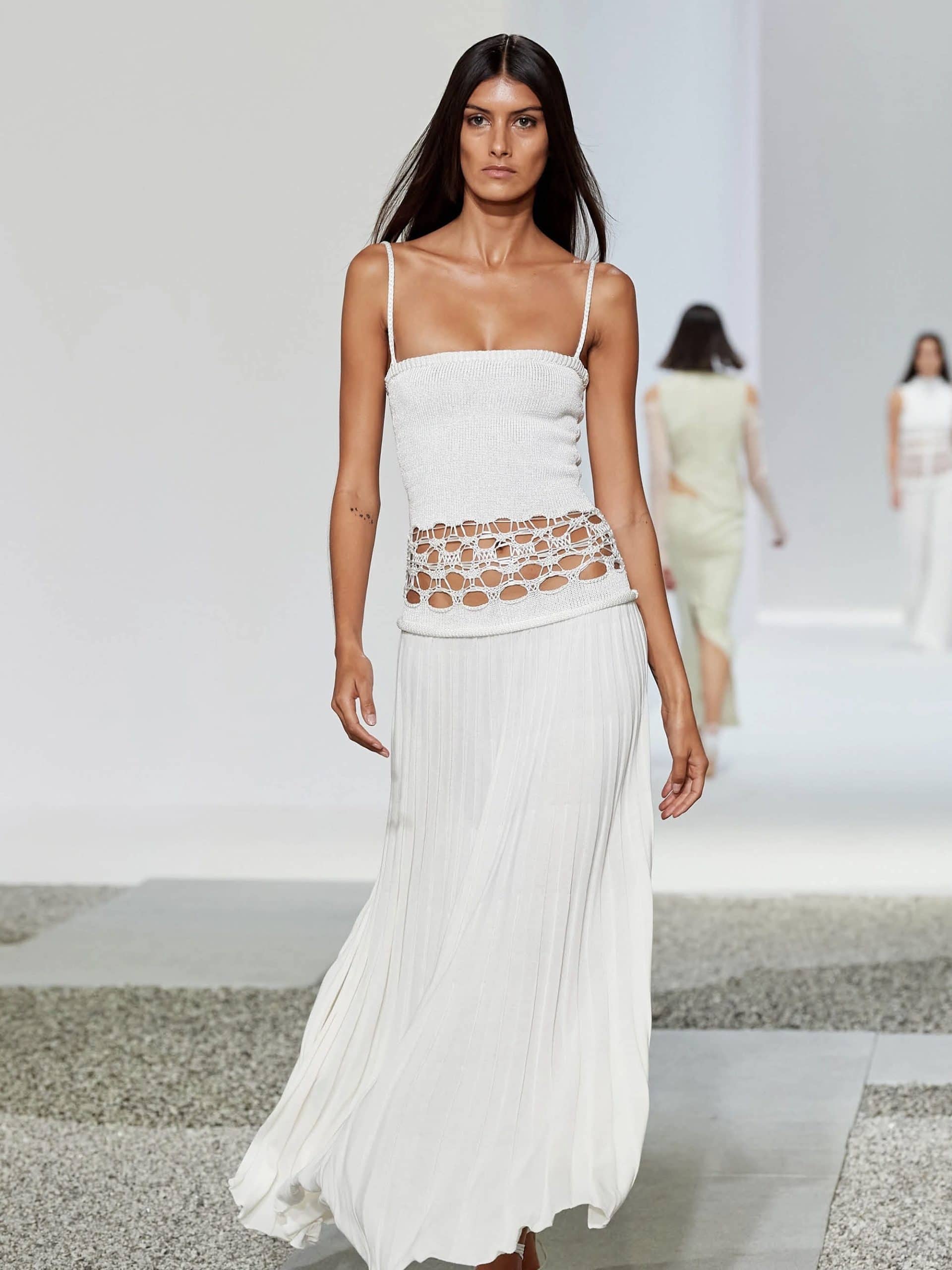 A female model walks on a runway wearing a white strappy top with a sheer, stone-embedded midsection and a flowing white skirt from the Pretty Eccentric Dresses That Are Bizarrely Fashionable collection. She has long brown hair and a serious expression.