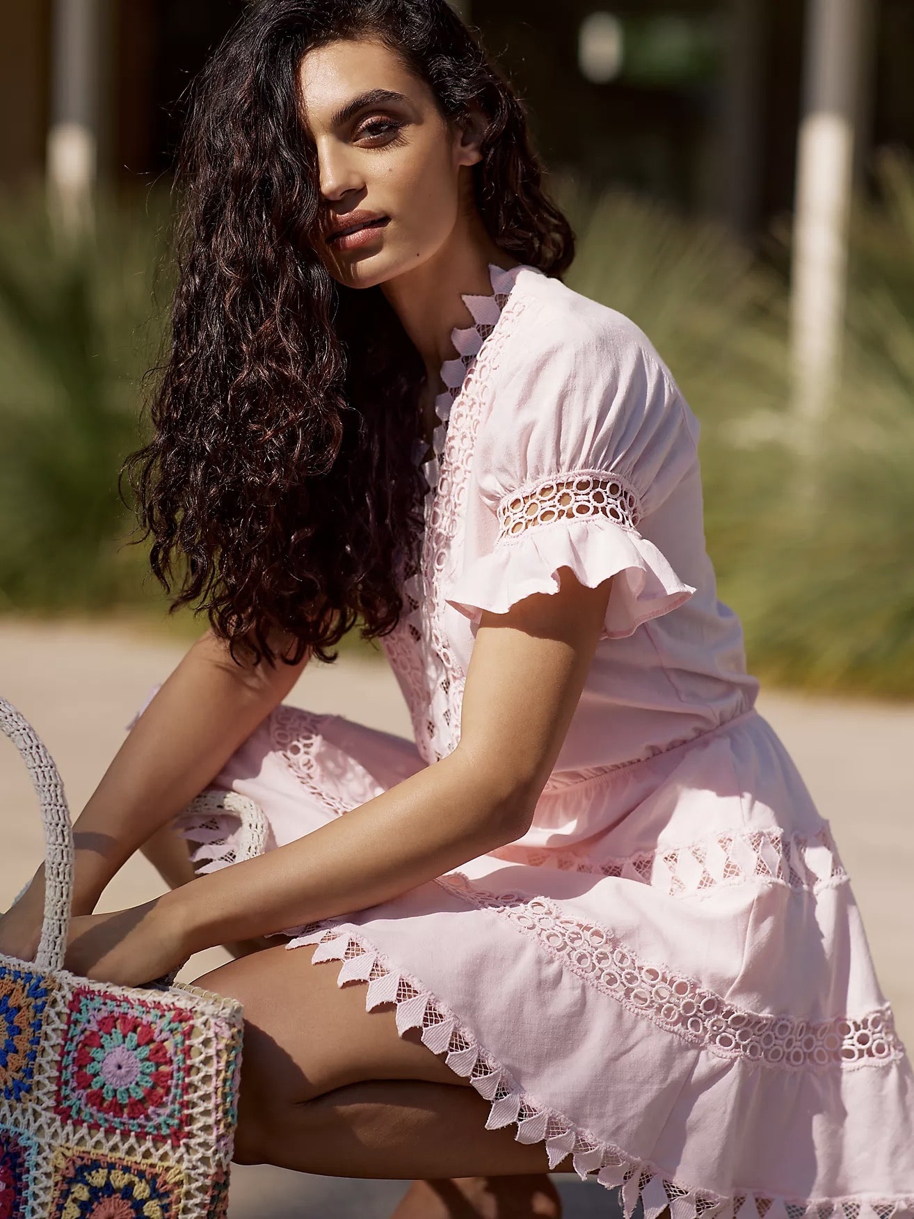 A woman with long curly hair crouches beside a colorful bag, wearing The Best Casual Dresses To Style With Sneakers with lace detailing, in a sunlit outdoor setting.