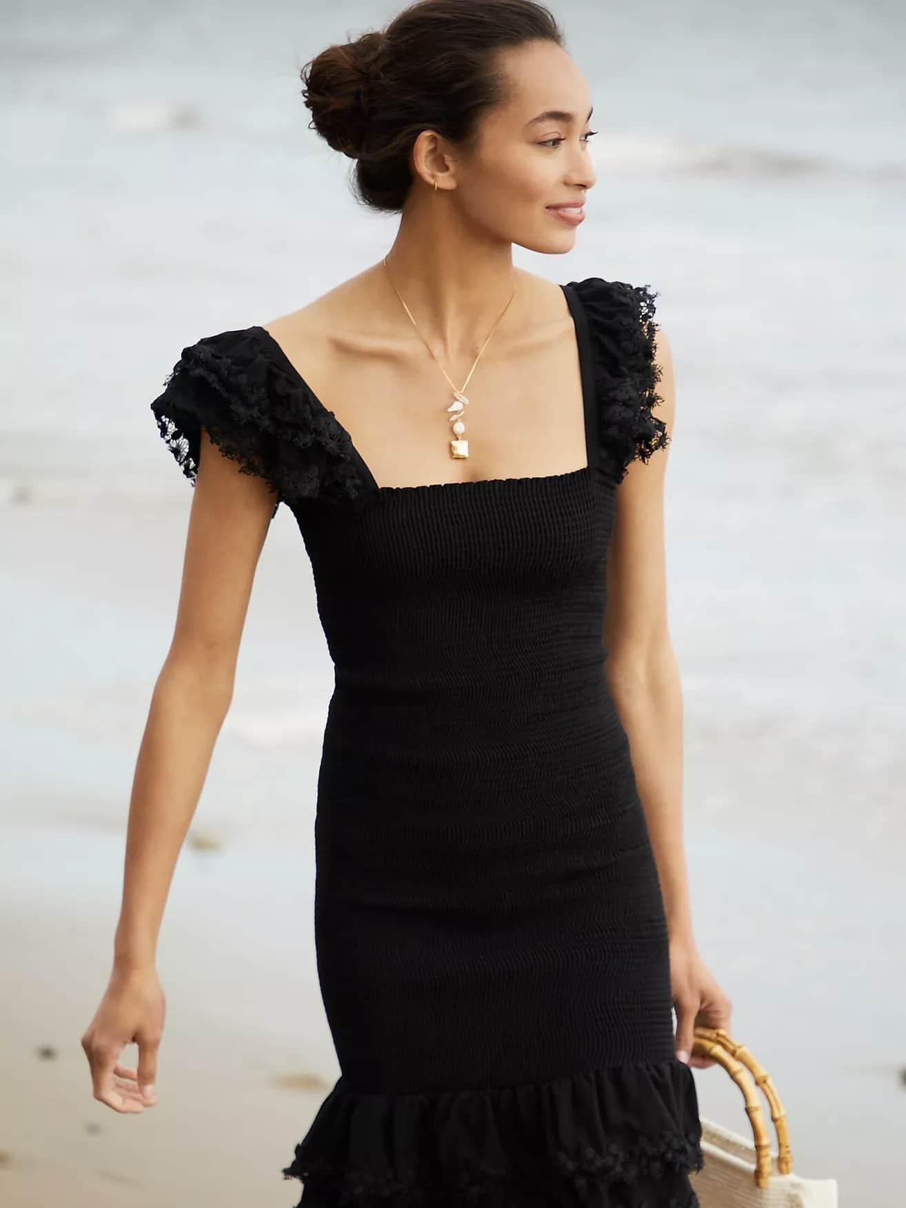 A woman in The Best Casual Dresses To Style With Sneakers and a necklace smiles gently while walking on a beach, carrying a straw tote bag. Her hair is styled in an elegant updo.