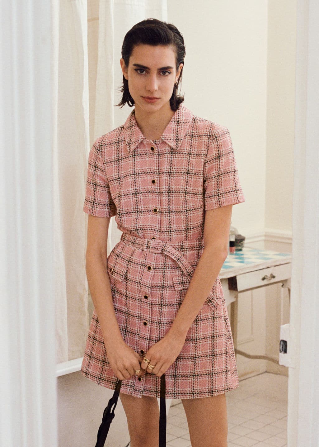 A person in a 30 Pink Designer Dresses You Need To Own stands in a bathroom, holding a handbag, with curtains partly open in the background. They have short dark hair and a serious expression.