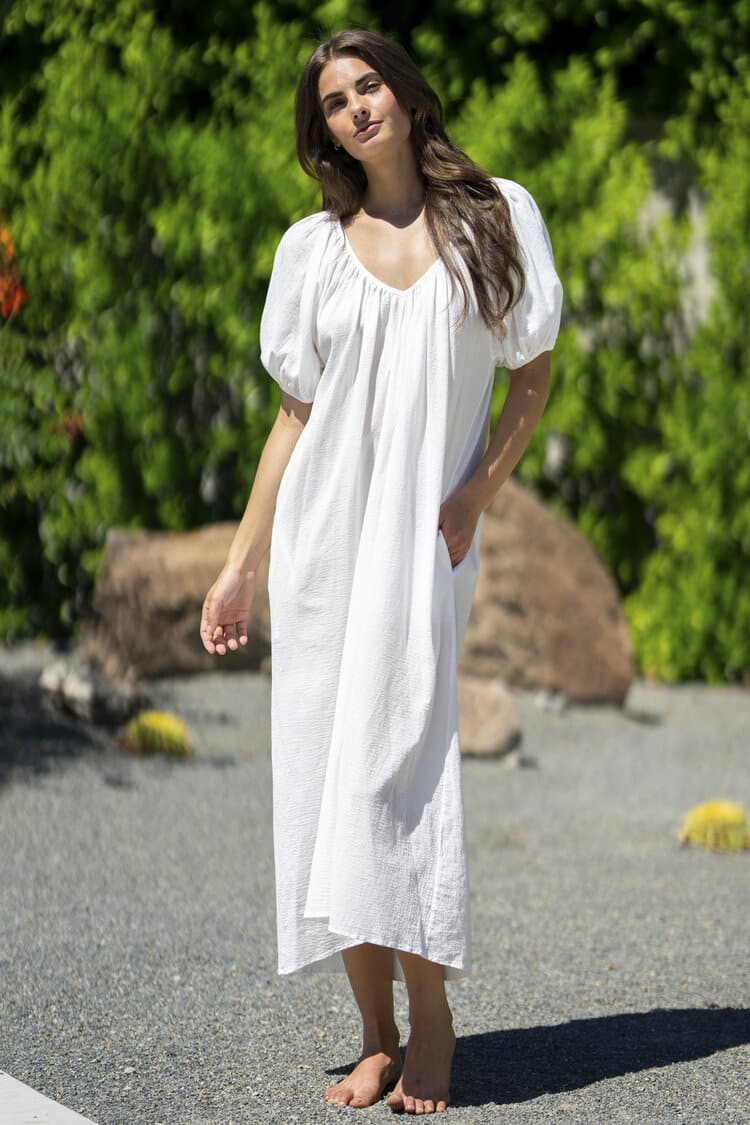 A woman in These Neutral Summer Dresses Are the Warm Weather Staples We Need stands barefoot on a sunlit pavement, surrounded by greenery and small yellow flowers, with a relaxed, contemplative expression.