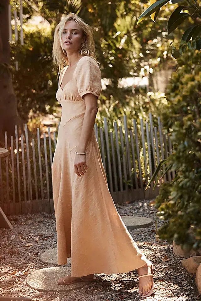 A woman stands in a sunlit garden, wearing These Neutral Summer Dresses Are The Warm Weather Staples We Need with short sleeves and a deep v-neckline. Her blonde hair is tousled, and she looks over her shoulder with