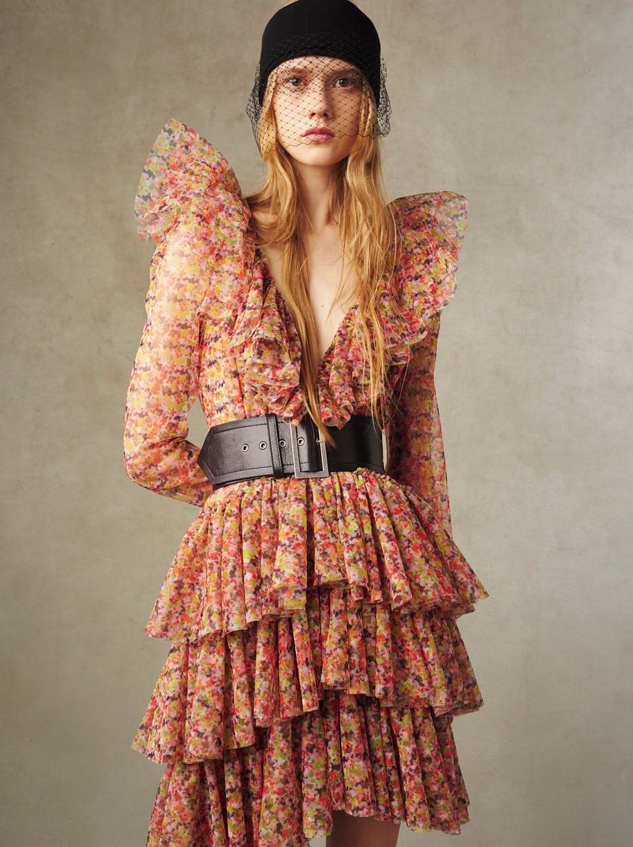 A woman models a Feel Playful And Pretty In Chic Short Tulle Dress with a wide black belt and a matching veiled hat. She stands against a neutral beige backdrop, her blonde hair flowing and her expression subtle.