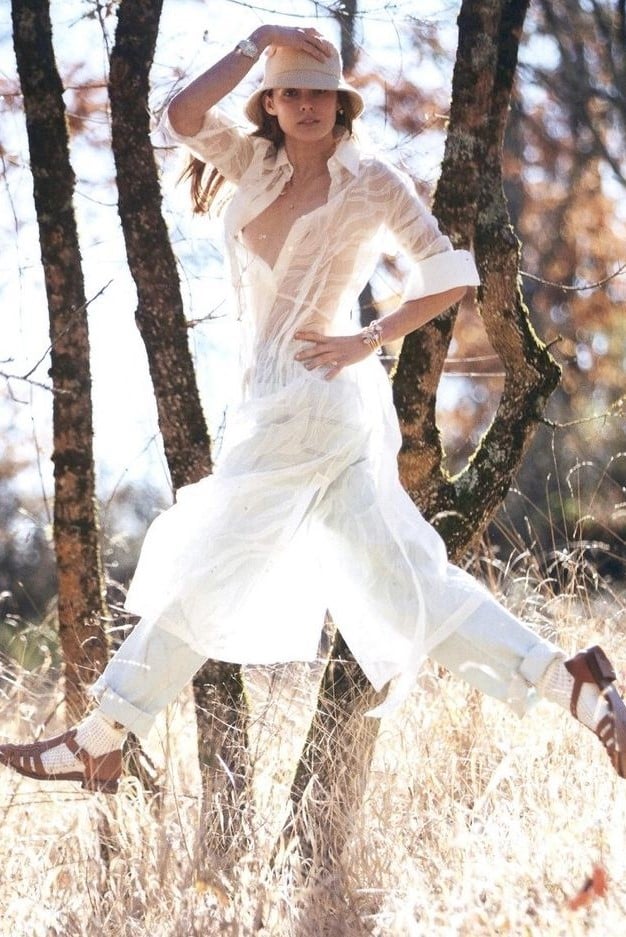 Sentence with product name: A woman in a flowing white dress and wide-brimmed hat is posing outdoors, leaning against a tree in a sunny, grassy field. She is wearing These Are The Perfect Designer Flats To Wear With Summer Dresses and has a serene expression.