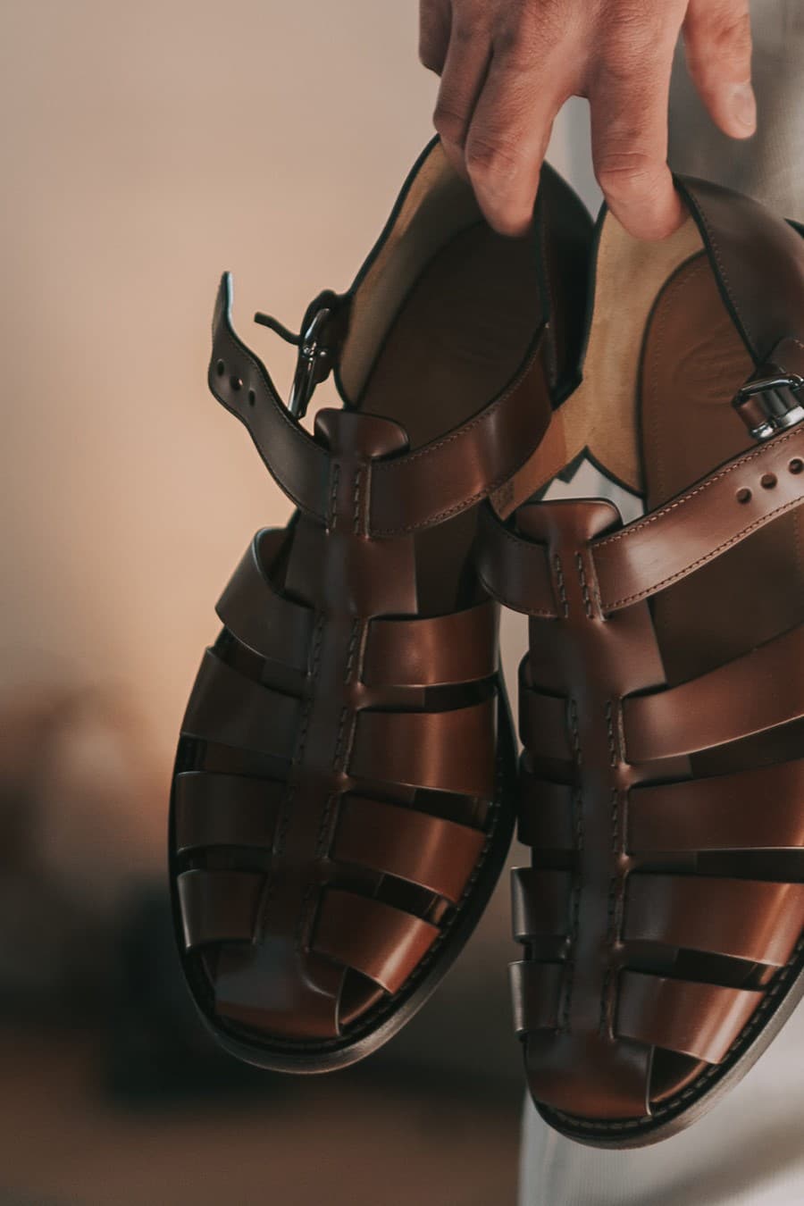 A person holding a pair of These Are The Perfect Designer Flats To Wear With Summer Dresses against a softly blurred background. The focus is on the sandals, showcasing their intricate straps and buckles.