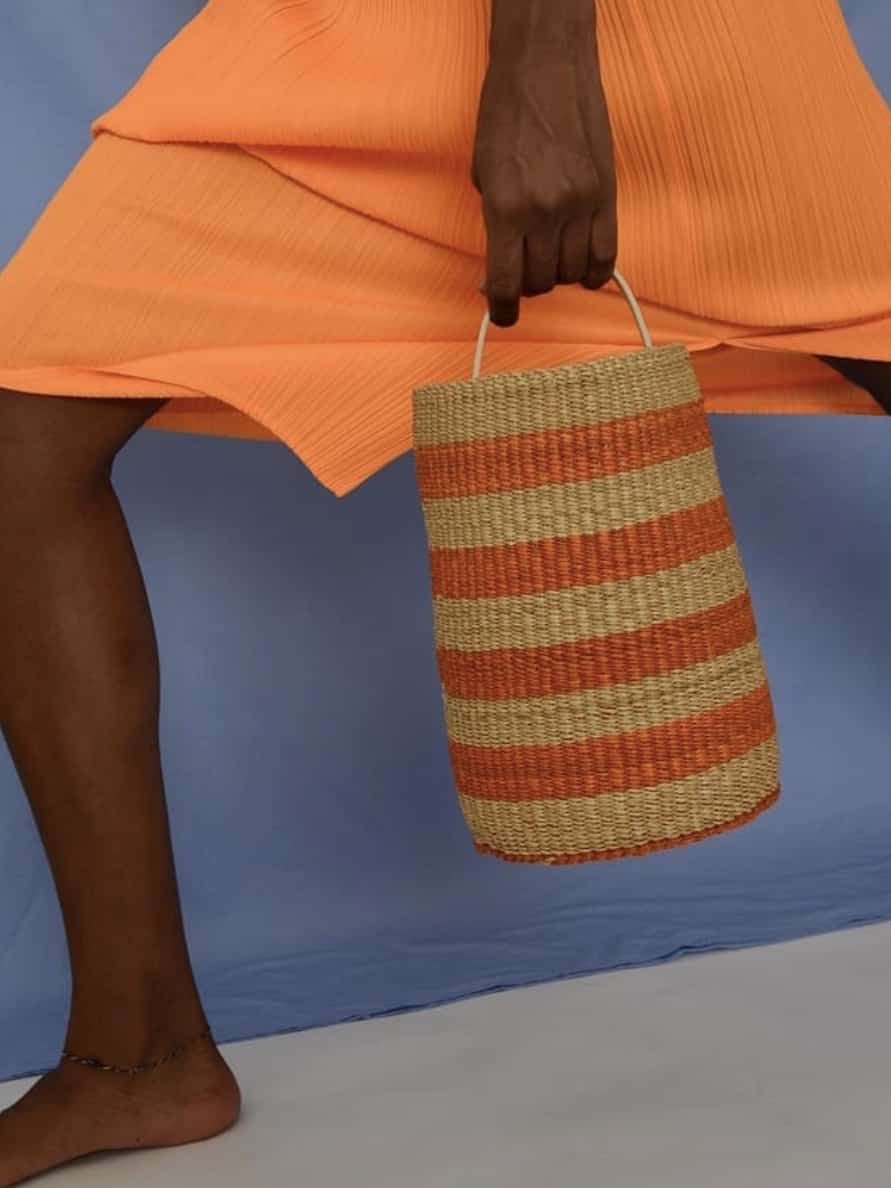 A person holding a striped Modern Beach Bags You Can Bring Off The Beach, standing in a pleated orange skirt against a blue background. Only the lower body from the waist down is visible.