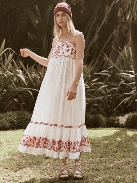 A woman in a white, floral-patterned "Boho Chic Dresses Are In Again…Here Are The Latest" dress and a pink beanie stands in a garden, holding out her hand slightly. She wears strappy sandals and looks directly at the camera.