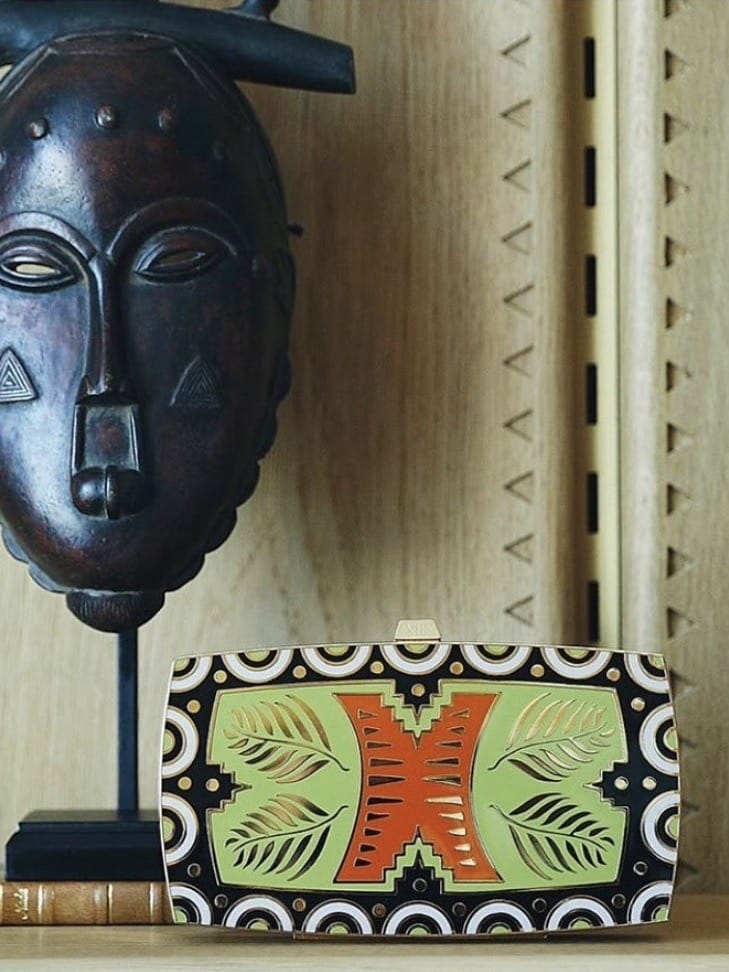 A wooden tribal mask displayed upright beside a Trendy Work Bag You Can Stylishly Wear To The Office on a wooden shelf, with a bamboo flute partially visible in the background.