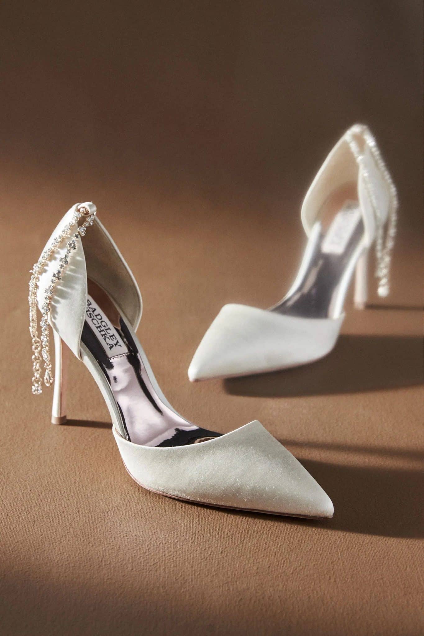 A pair of elegant ivory "We've Found All The Most Perfect Bridal Shower Heels" with pointed toes and decorative pearl chains draped over one shoe, set against a neutral beige background.