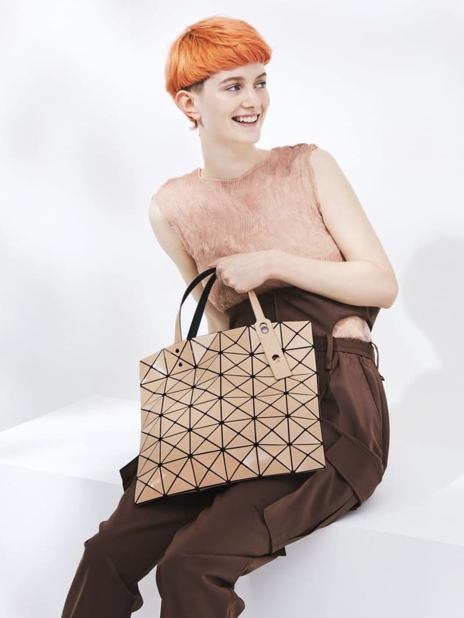 A young woman with short orange hair, smiling and sitting. She wears a sleeveless top and brown trousers, holding a large, Trendy Work Bags You Can Stylishly Wear To The Office.