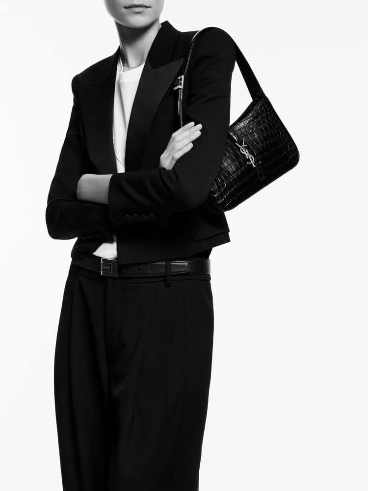 A monochrome image of a person dressed in a tailored black blazer and trousers, arms crossed. The person is holding a trendy Work Bags You Can Stylishly Wear To the Office shoulder bag. Only the torso and arms are visible.