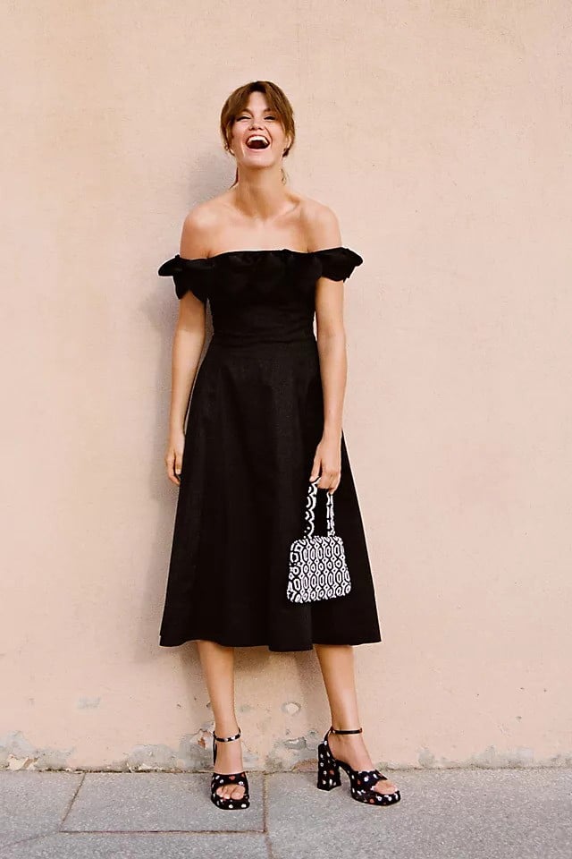A woman in a You’ll Want To Own Every One Of These beautiful black dress and polka dot heels, laughing joyfully with her head tilted back, standing against a plain beige wall while holding a patterned handbag.