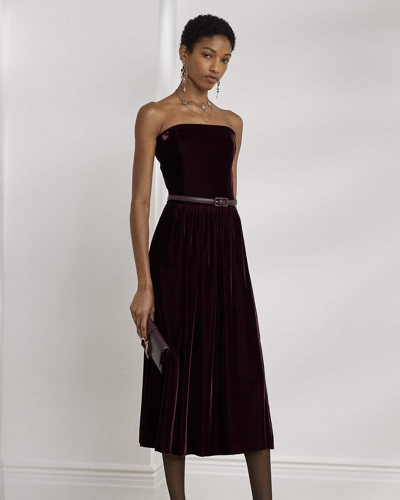 A woman in a dark maroon strapless velvet midi dress from These Capsule Wardrobe Dresses Will Never Go Out Of Style, with a thin belt, holds a small clutch and stands against a plain white background. She wears dangling earrings and looks