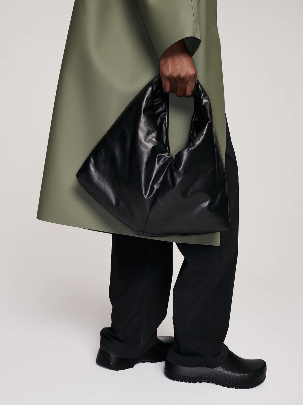 A person in a green coat and black trousers holds a glossy [Product Name] Large Size Bag. Only the lower half of their body is visible. They wear chunky black shoes.