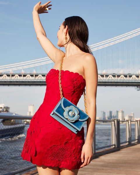 A woman in a red iconic day dress and holding a blue purse stands by a riverside, gesturing skyward with her hand, with a bridge and city skyline behind her, wearing an Iconic Day Dress To Be Seen In At New York Fashion Week.