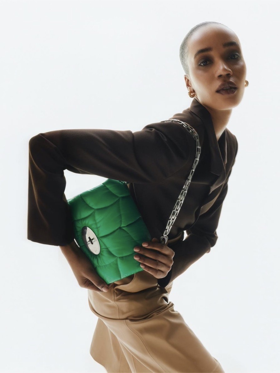 A woman with a shaved head, wearing a dark blazer and tan pants, poses holding a Large Green Minimalist Designer Bag with a silver chain strap. The background is a light, neutral color.