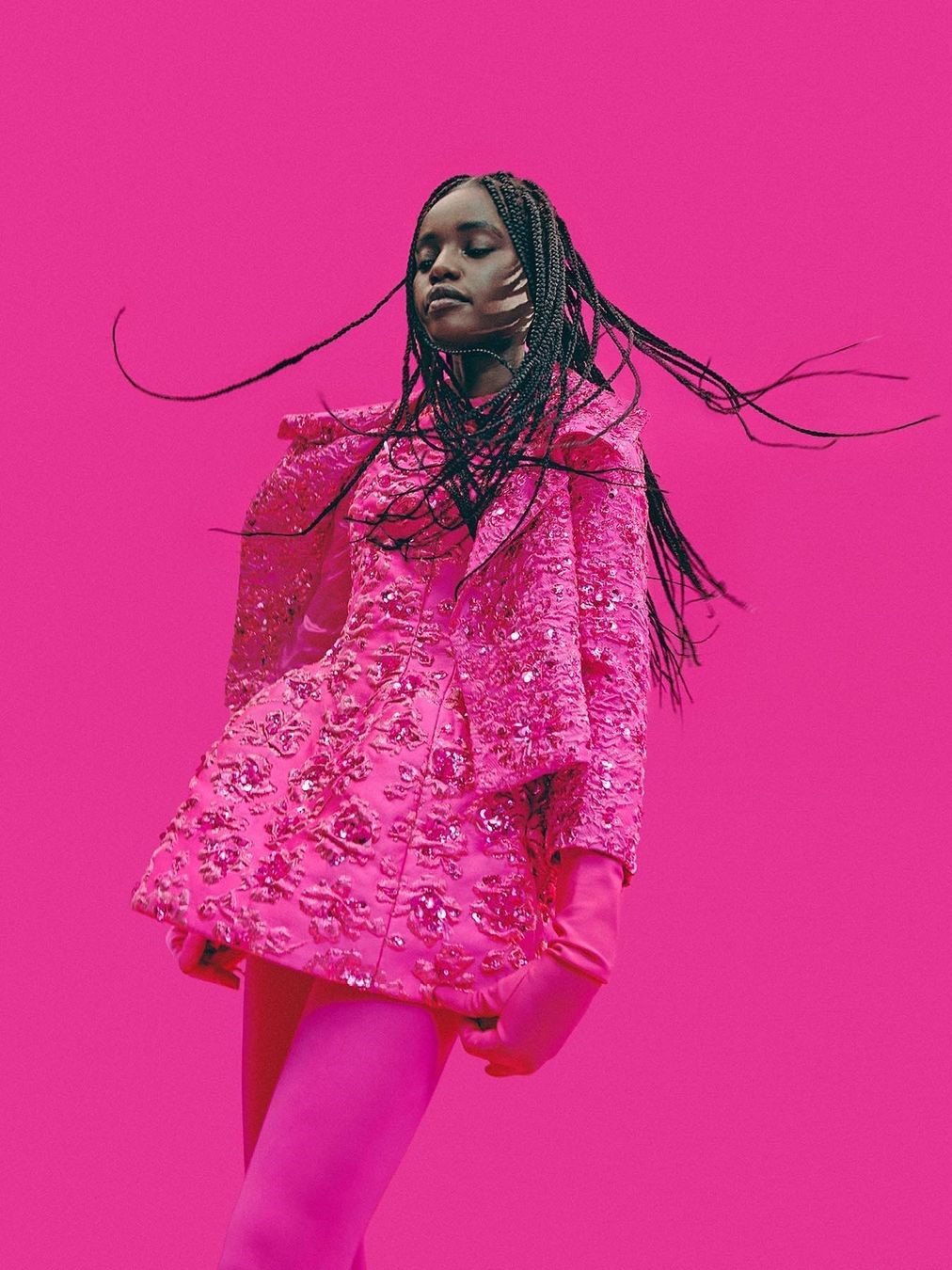 A woman in a sequined Feel Fierce In Trending Fuchsia Color Dresses jacket and matching pants stands against a pink background, her long braided hair flowing dynamically around her.
