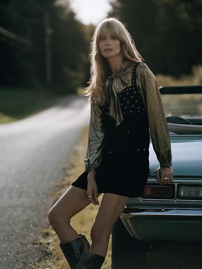 A woman with blonde hair leans against a classic car on a quiet road surrounded by trees, dressed in These Velvet Party Dresses Will Be Your Best Investment This Season, star-patterned scarf, and tall boots.