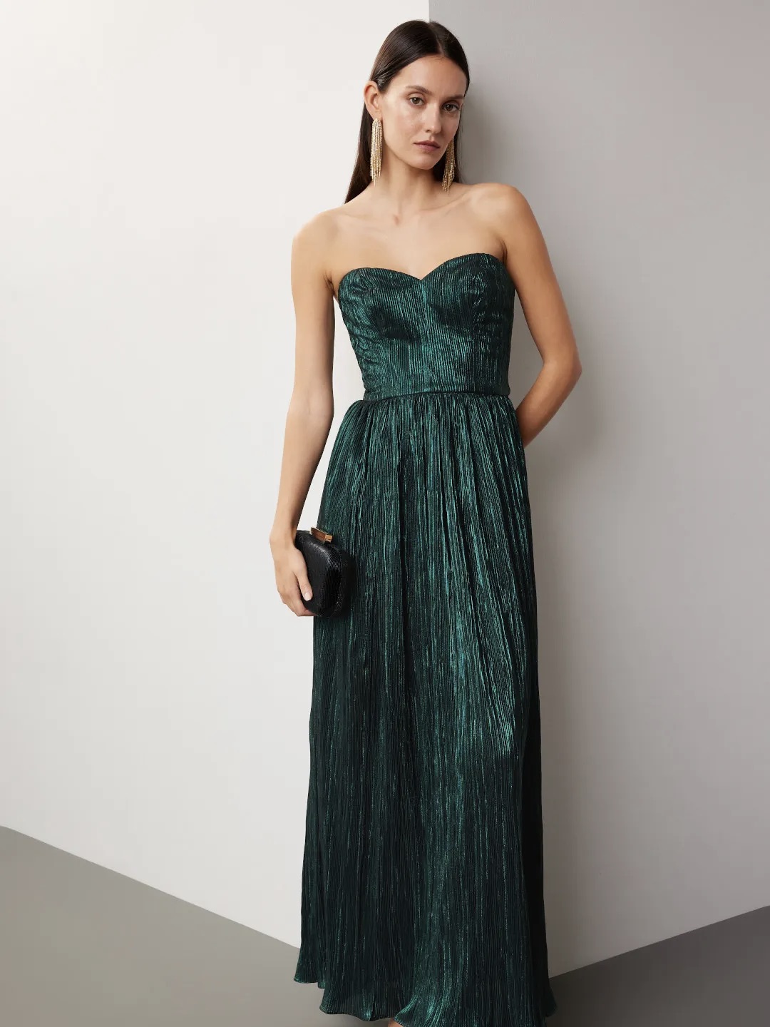 A woman in a DRESS THE POPULATION Audrina Strapless Gown stands against a gray background, holding a black clutch, with her hair pulled back and a focused expression.