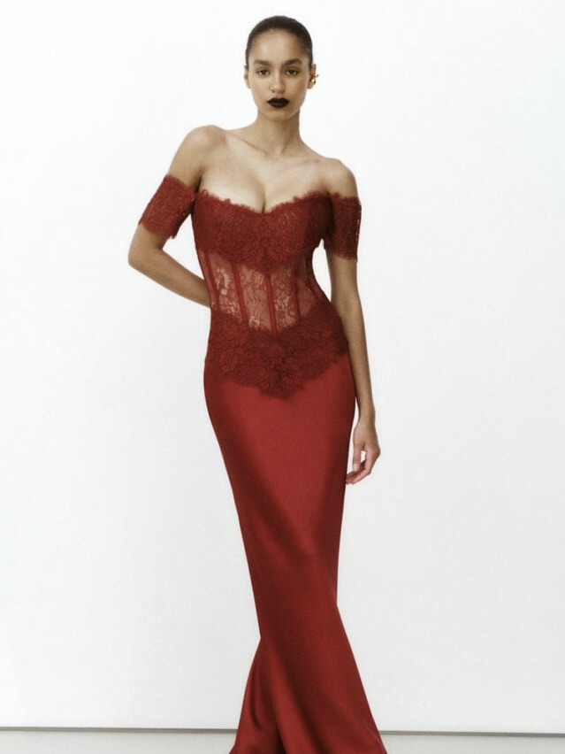 A woman models an elegant red evening gown suitable for black tie events, with an off-the-shoulder lace bodice and a fitted, floor-length skirt. She stands against a plain white background in the "From Weddings To Holiday Galas—These Black Tie Dresses Will Take Your Breath Away.