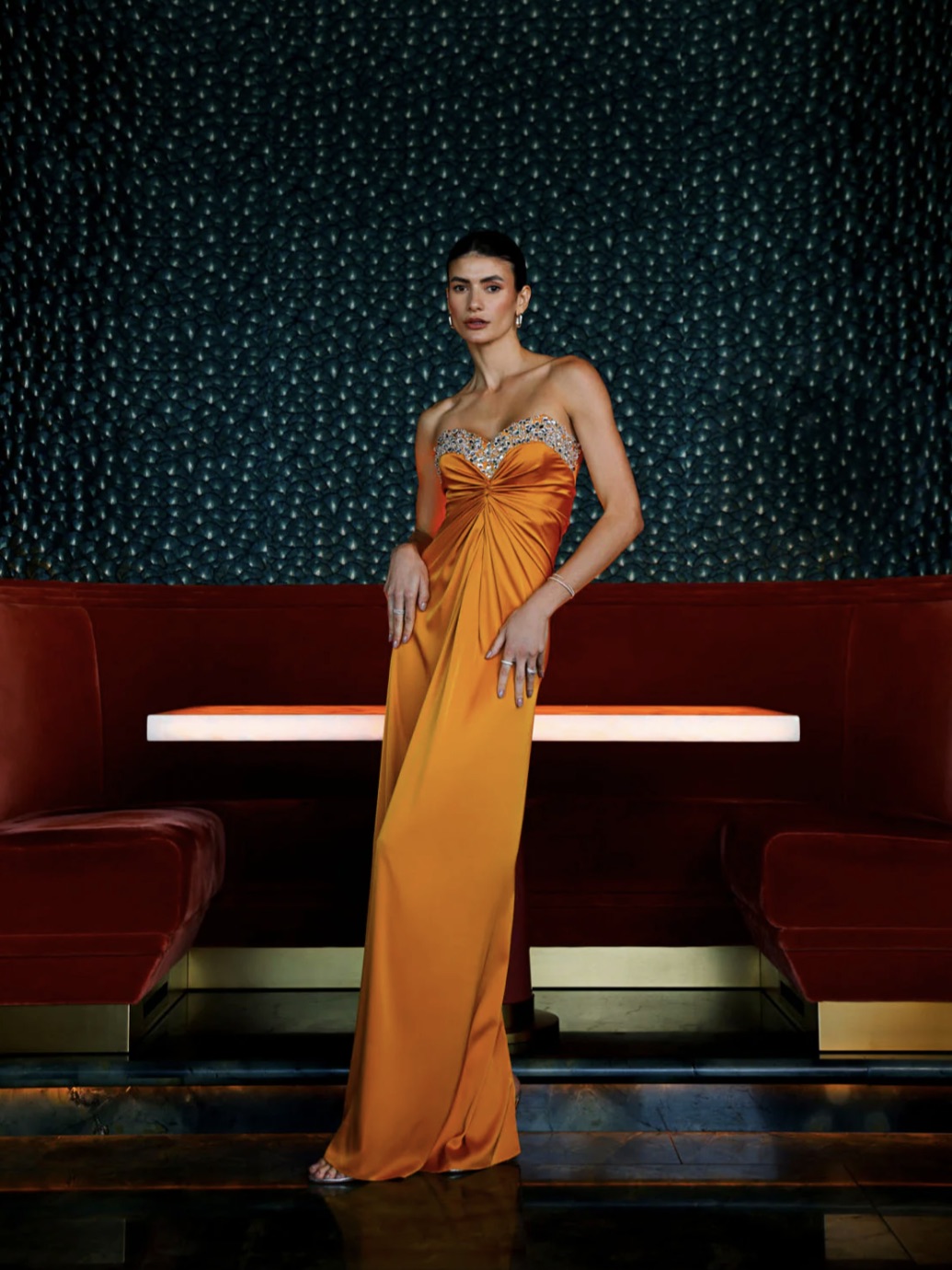 A woman in an elegant orange gown fit for From Weddings To Holiday Galas—These Black Tie Dresses Will Take Your Breath Away stands in a luxurious room with red velvet sofas and a shimmering blue backdrop. Her posture exudes confidence as she gazes directly at the
