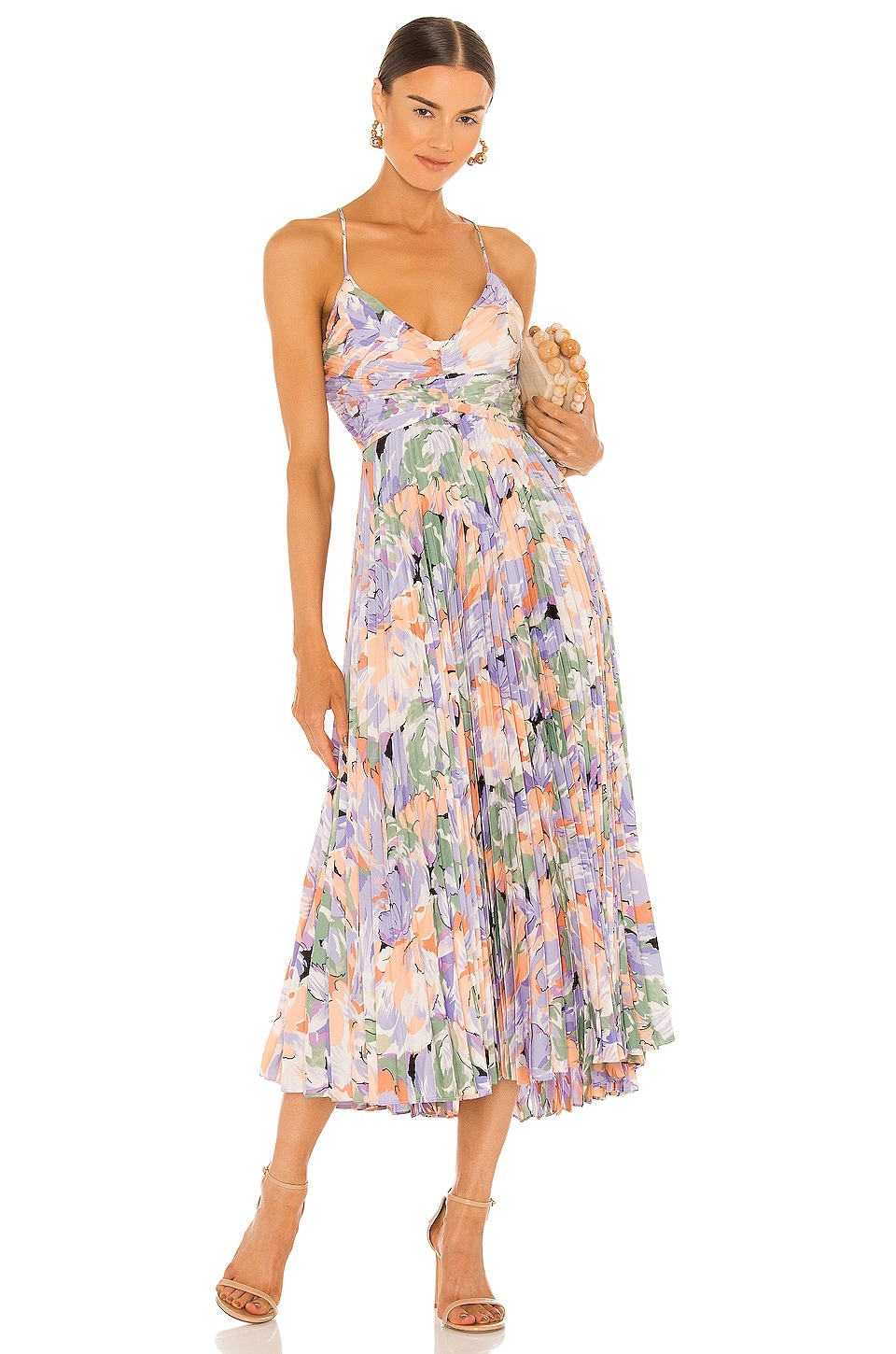 A woman wearing an ASTR THE LABEL Blythe Dress 1 floral pleated midi dress with thin straps, paired with beige high-heeled sandals, standing against a white background. She accessorizes with a small clutch and drop earrings.