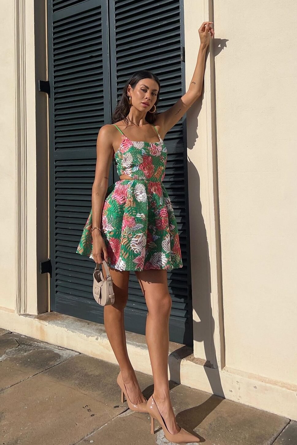 A woman in a {Printed Designer Mini Dress We’re Bookmarking For Spring & Summer} and heels stands by a building with shutters, holding a handbag, with sunlight casting shadows on the wall.