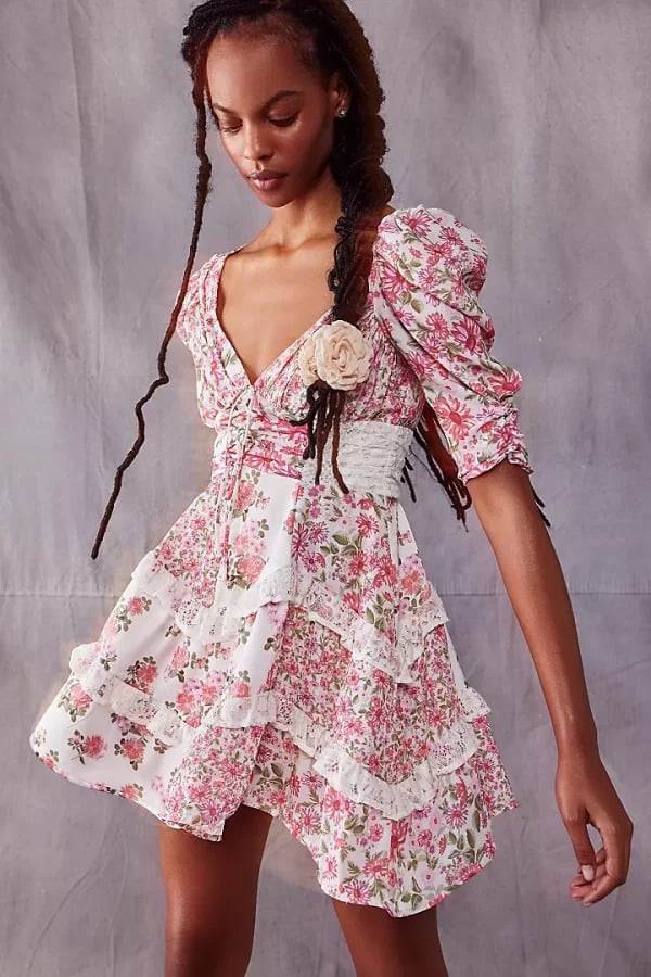 A woman in a Printed Designer Mini Dress We're Bookmarking for Spring & Summer with puffed sleeves and a lace-up corset waist stands against a grey background. She has long, braided hair and holds a soft, contemplative pose.