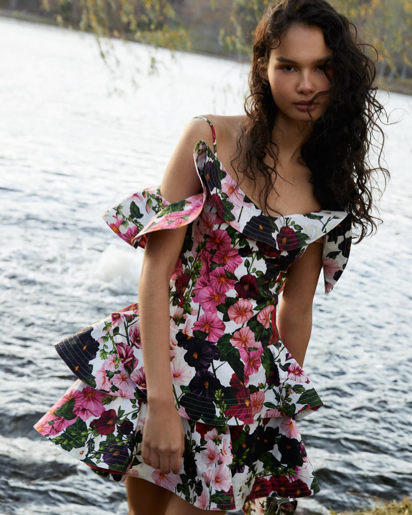 A young woman wearing a Printed Designer Mini Dress stands by a lakeside, her curly hair tousled. The background features tranquil water and sparse foliage.