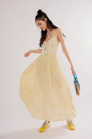 A woman in a FREEPEOPLE Sweet Nothings Midi Dress paired with yellow sneakers, holding a colorful handbag, looks downwards while striking a playful pose against a light background.