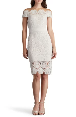 A woman is standing wearing an off-the-shoulder white TADASHI SHOJI Corded Lace Sheath Cocktail Dress that falls to the knee, paired with light-colored strappy heels.