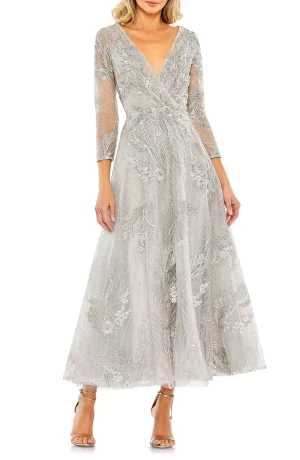 A woman is wearing a silver, ankle-length MAC DUGGAL Embellished Faux Wrap A-Line Cocktail Dress with long, sheer sleeves and a deep V-neckline. The dress features intricate floral embroidery and is paired with metallic high-heeled sandals. She stands against a plain white background.