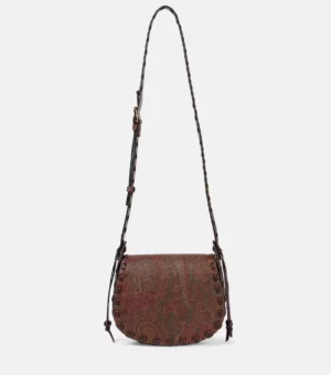 The Etro Arnica 1984 Drya Small Crossbody Bag features a brown paisley design, adjustable strap, rounded flap, decorative stitching along the edges, and small hanging tassels on both sides.