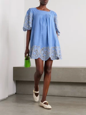 A person wearing a loose light blue Thierry Colson Scarlett Broderie Anglaise Linen and Cotton-blend Mini Dress, cream woven shoes, and holding a small green handbag, stands indoors against a neutral background.