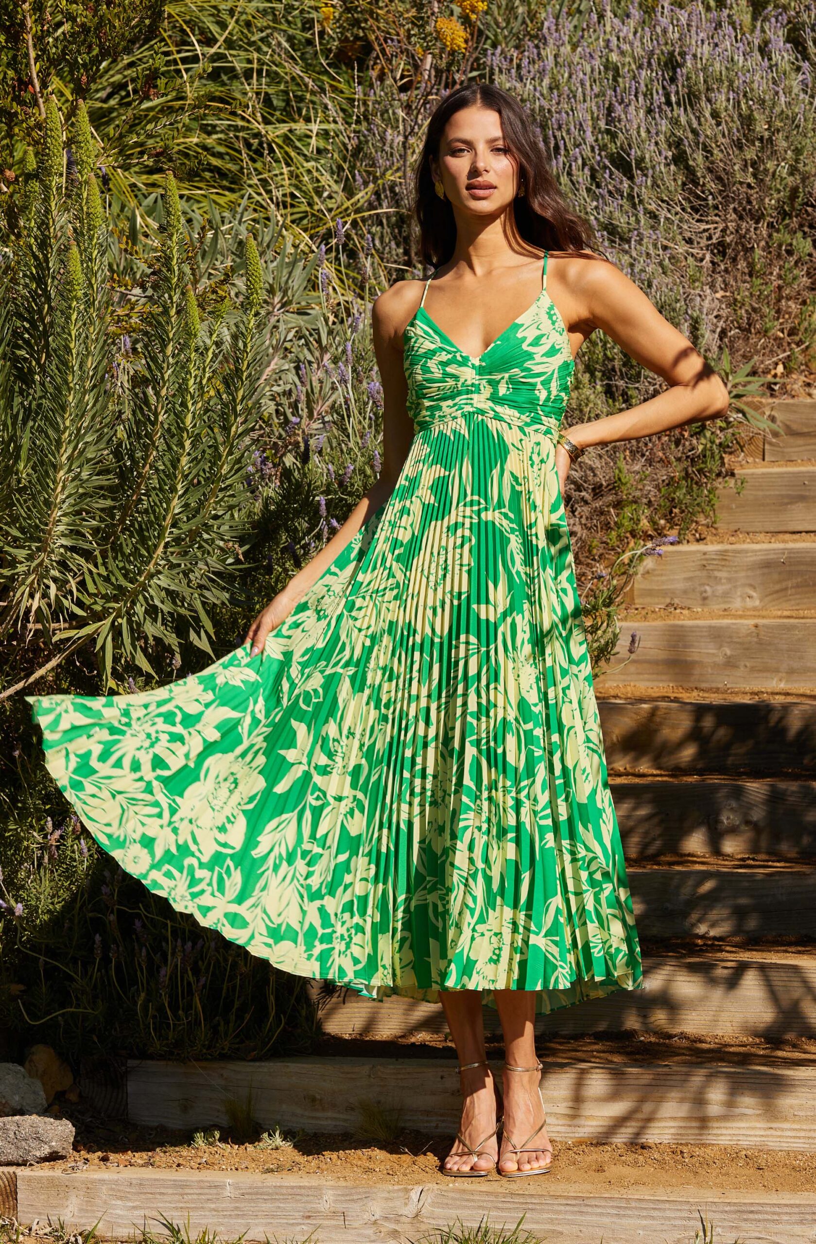 A woman stands outdoors on wooden steps, wearing the ASTR THE LABEL Blythe Dress, a green and yellow floral pleated midi with thin straps. She holds out one side of the skirt, posing with one hand on her hip amid lush greenery in the background.
