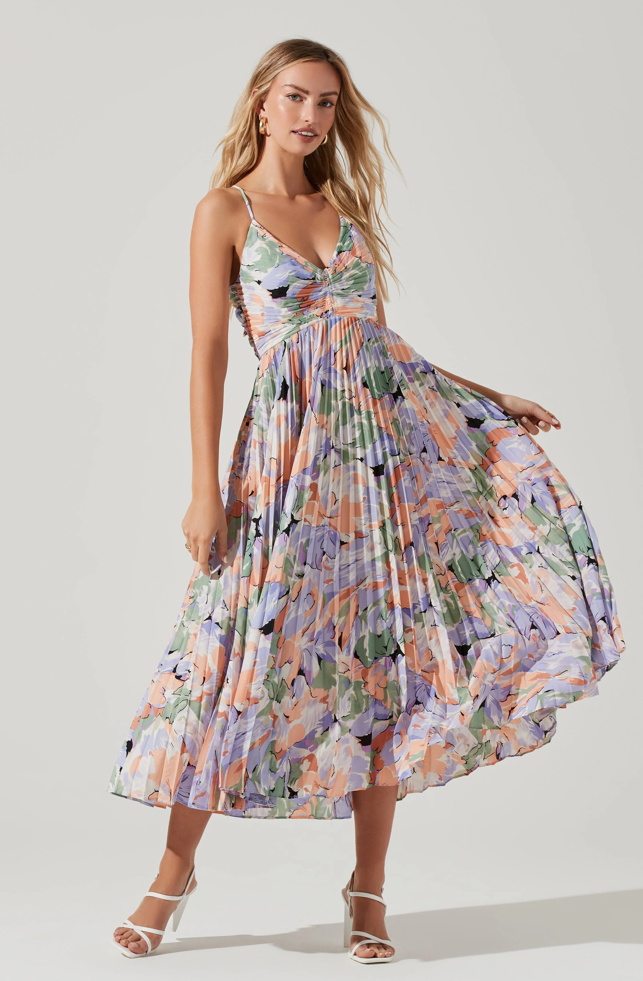 A woman wearing the ASTR THE LABEL Blythe Dress—a sleeveless, V-neck, pleated midi with a pastel floral pattern—stands against a plain background. She holds one side of the dress out and pairs it with white heeled sandals.