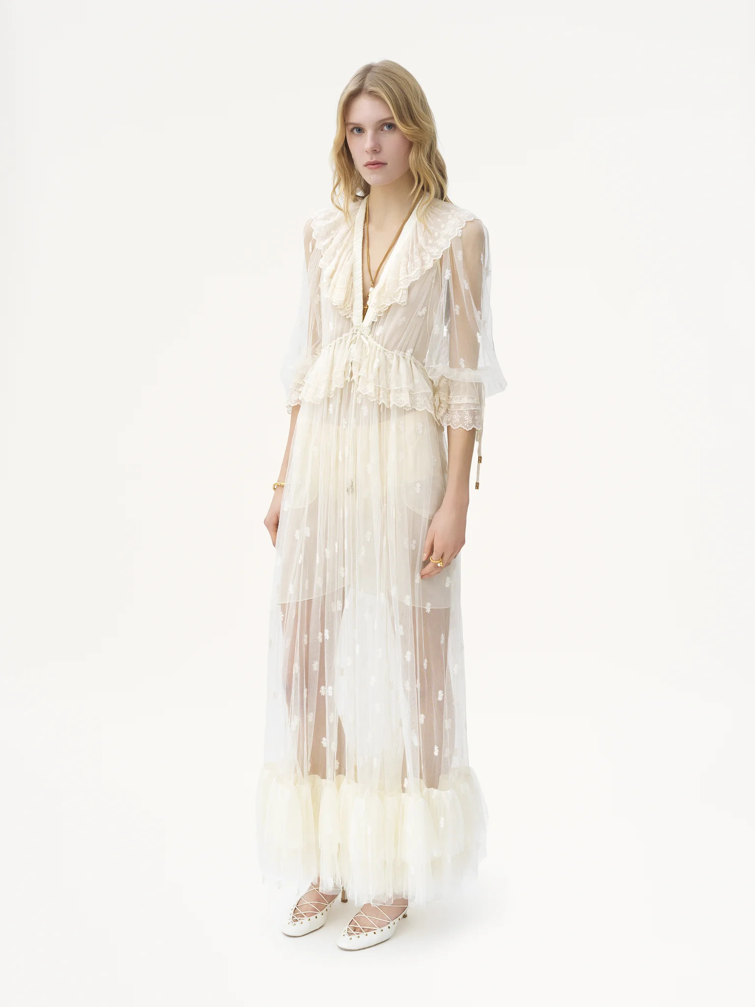 A person stands against a plain white background in a long, sheer, cream-colored dress with ruffles and star patterns—a whimsical look inspired by 2025 wedding dress trends, featuring a large collar and feathered trim at the hem.