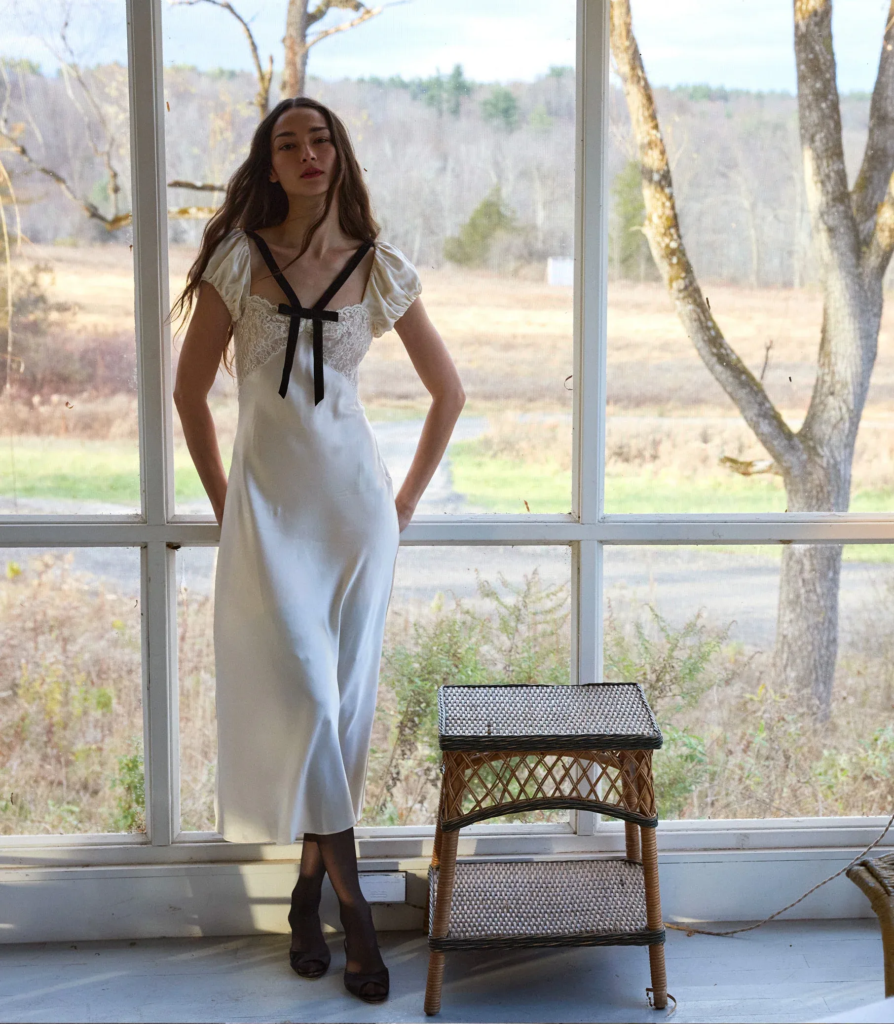 A woman in a long white wedding dress with black ribbon detail stands indoors by a window. She has long hair and her hands in her pockets—a modern nod to wedding dress trends for brides 2025. Bare trees and open fields are visible outside.