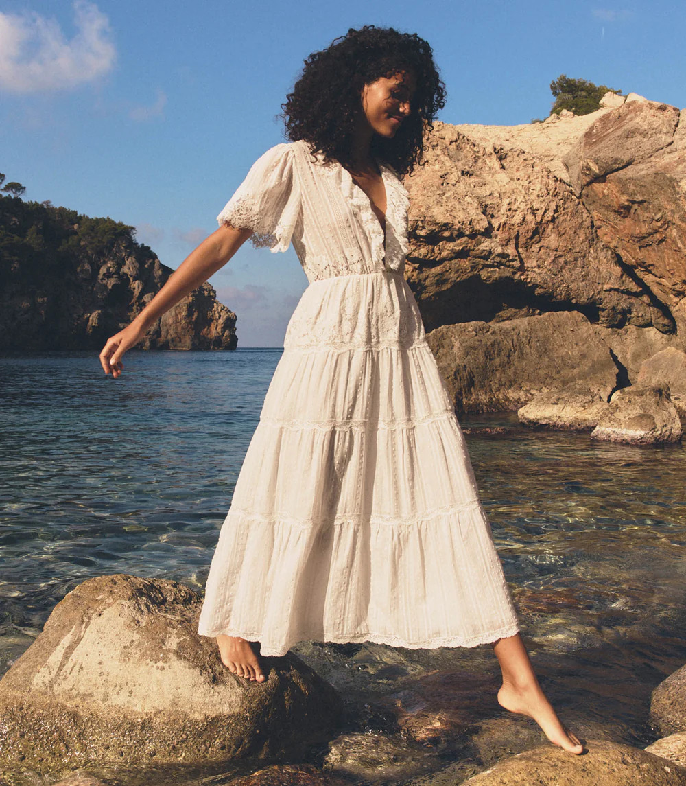 A woman in a white, short-sleeved, ankle-length dress stands barefoot on rocks by clear blue water—an elegant vision of white dresses set against rocky cliffs and lush greenery beneath a partly cloudy sky.