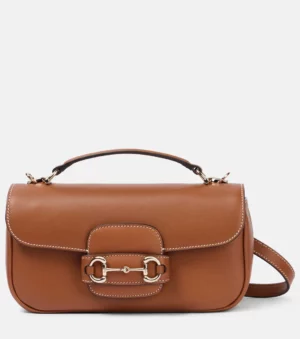 A brown leather tote bag with white stitching, a top handle, front flap, gold hardware, and a decorative buckle detail inspired by the Horsebit 1955. Features a removable shoulder strap for versatile wear.