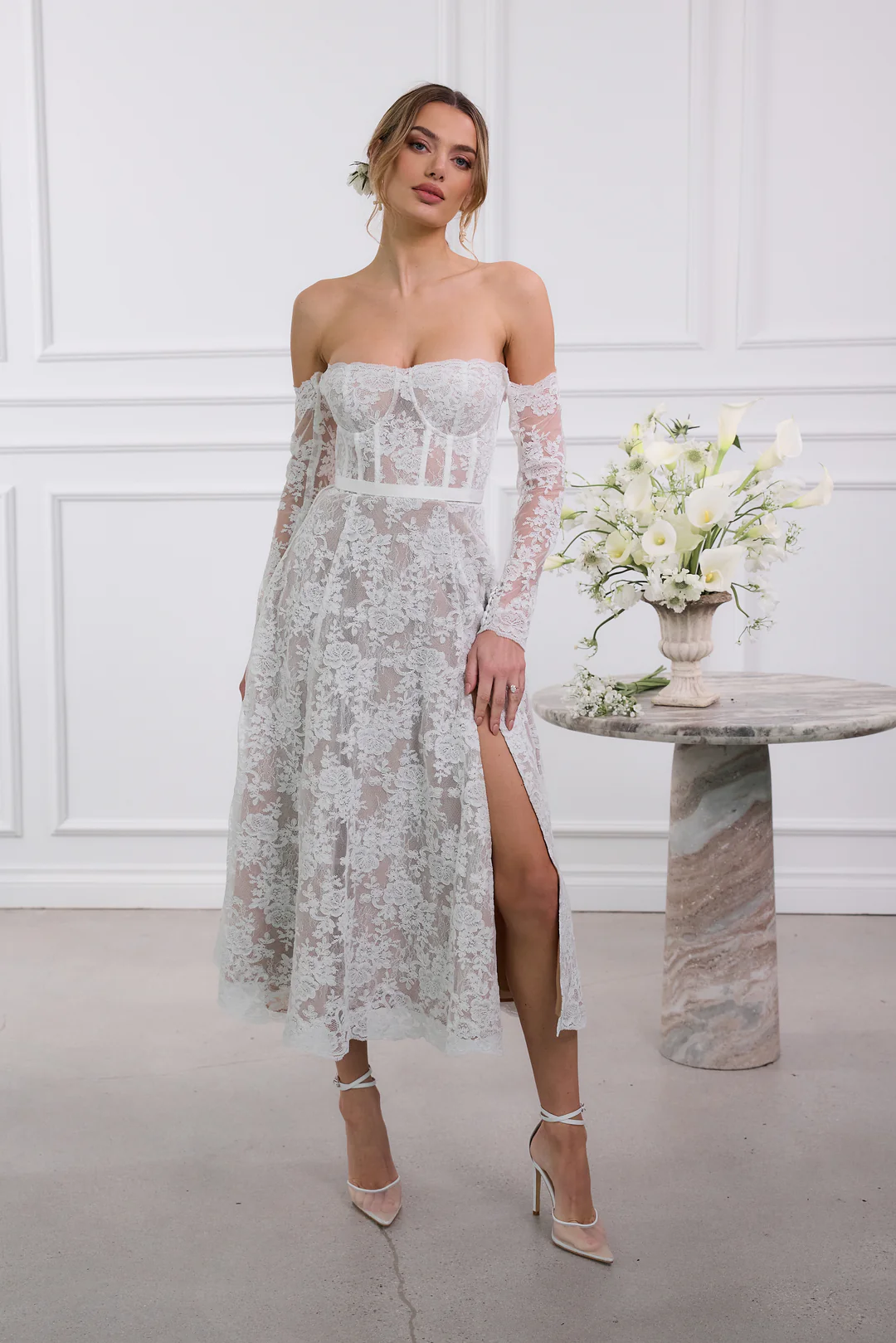 A woman wearing an off-the-shoulder, long-sleeved, white lace wedding dress with a corset bodice and thigh-high slit—perfect for brides following 2025 wedding dress trends—stands indoors beside a round stone table topped with a white floral arrangement.