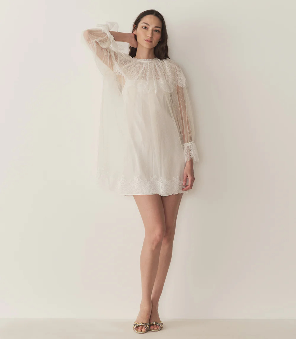 A woman stands against a plain white background wearing a short, sheer white dress with lace details, long sleeves, and ruffles. This stylish pick from our White Dresses collection is paired with open-toed sandals and her long dark hair flows freely.