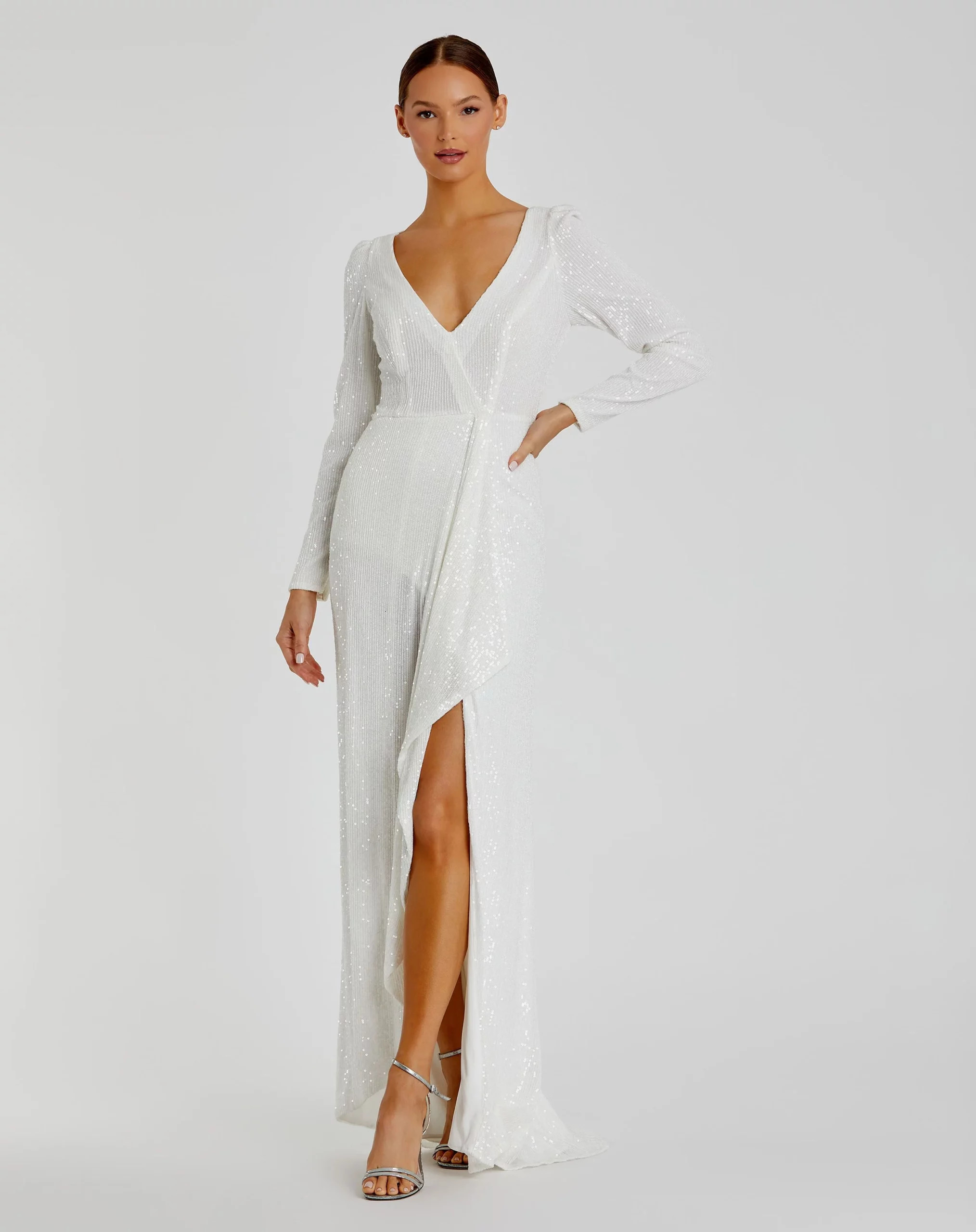 A woman stands against a plain background in a long-sleeve, white sequin wrap dress with a deep V-neckline and high front slit—an elegant choice for brides seeking wedding dress trends for 2025. She poses confidently in silver high-heeled sandals.