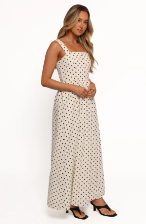 A woman stands against a plain white background wearing the Jenni Polka Dot Maxi Dress—sleeveless, ankle-length, with black polka dots and black heeled sandals. She has long, wavy hair and a neutral expression.