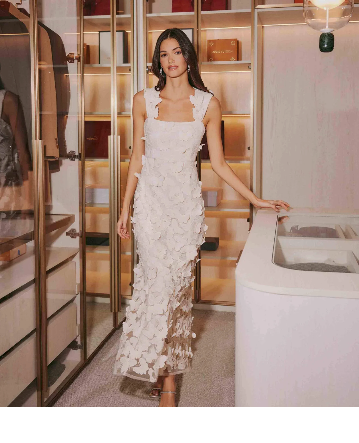 A woman stands in a modern walk-in closet wearing a sleeveless, ankle-length white dress with floral appliqués—an elegant nod to wedding dress trends for brides 2025. She has long, straight hair and rests one hand on a white counter.