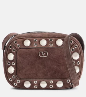 A brown Nellcote Small Suede Crossbody Bag with silver studs and grommets, featuring a zipper closure, adjustable strap, and a small metal Valentino logo at the center front.