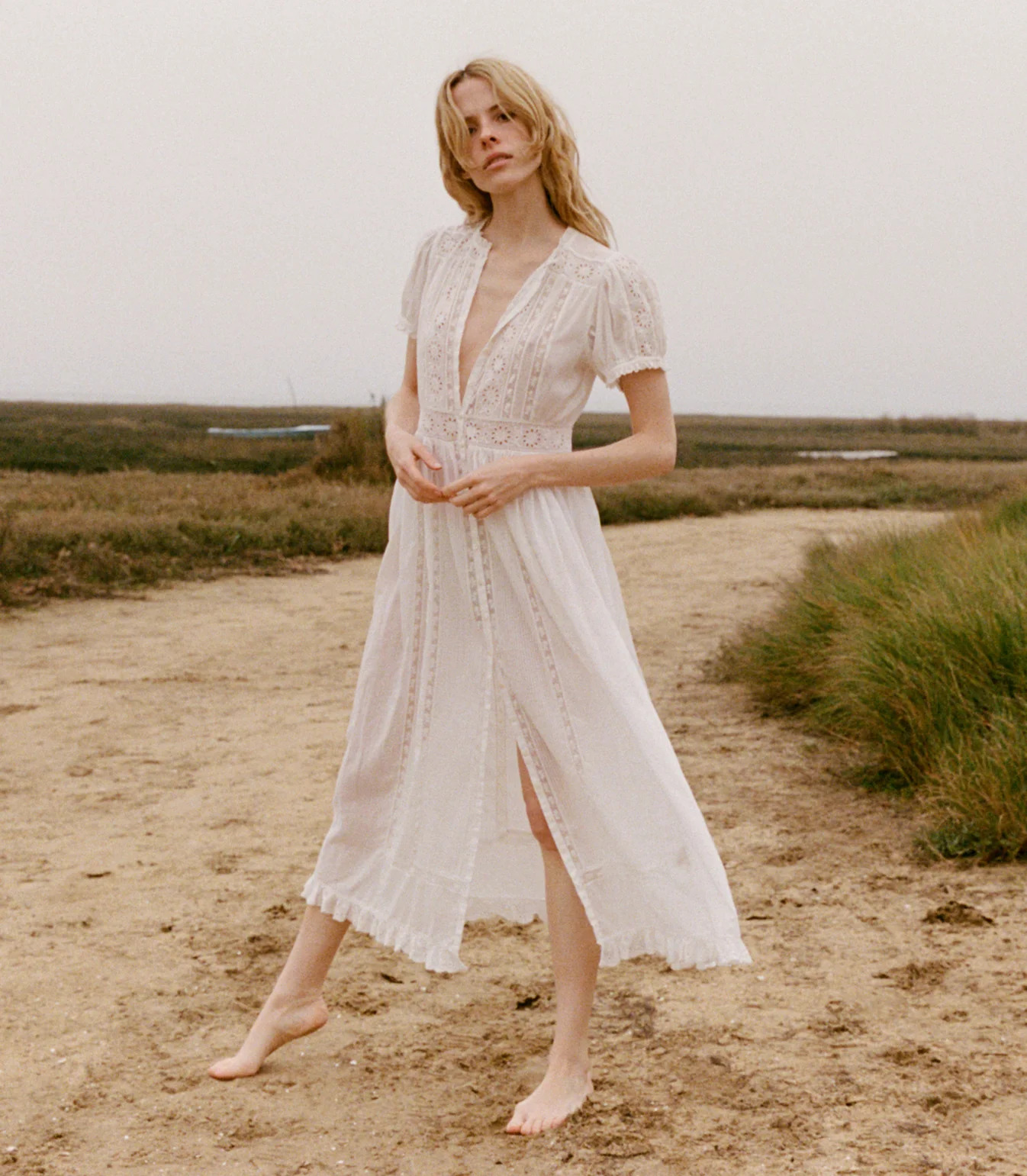 A barefoot woman in a lace-trimmed white dress, perfect for lovers of white dresses, stands on a dirt path in a grassy, rural area beneath an overcast sky.
