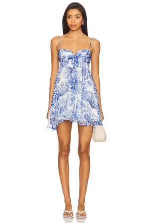 A woman stands facing forward in the Amanda Uprichard Liana Dress, a short blue and white leaf-patterned style with thin straps and ruffle details. She accessorizes with a small white handbag and white sandals.