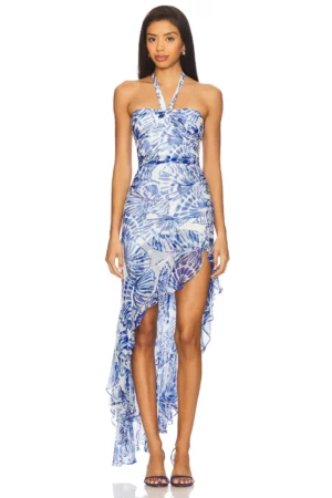 A woman stands facing forward wearing the Amanda Uprichard Liana Dress, a strapless blue and white halter style with a leaf pattern and asymmetrical ruffled hem, paired with open-toed heels.