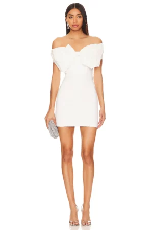 A woman stands facing forward wearing a short, off-the-shoulder Bardot Triple Frill Dress with a large bow detail on the chest. She holds a silver clutch and wears clear high-heeled sandals.